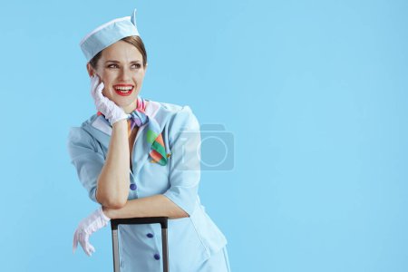 Foto de Asistente de vuelo femenino elegante feliz aislado sobre fondo azul en uniforme azul con bolsa de carro mirando espacio de copia. - Imagen libre de derechos