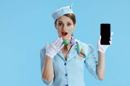 Téléchargez les photos : Surprise hôtesse de l'air féminine moderne sur fond bleu uniforme bleu montrant écran vide smartphone. - en image libre de droit