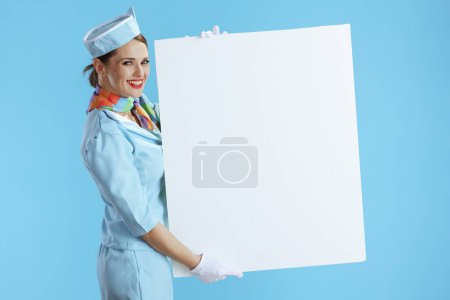 Photo for Happy modern female flight attendant against blue background in blue uniform showing blank board. - Royalty Free Image