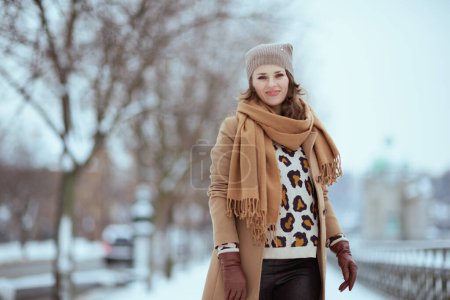 Téléchargez les photos : Heureuse femme élégante en chapeau marron et écharpe en manteau de chameau avec des gants à l'extérieur de la ville en hiver. - en image libre de droit