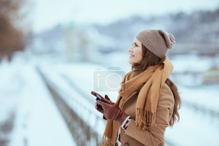 Photo for Smiling elegant woman in brown hat and scarf in camel coat with gloves sending text message using smartphone outside in the city in winter. - Royalty Free Image