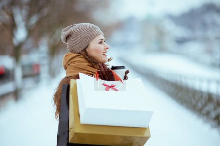 Foto de Sonriente mujer con estilo en sombrero marrón y bufanda con guantes, bolsas de compras y taza de chocolate caliente al aire libre en la ciudad en invierno. - Imagen libre de derechos