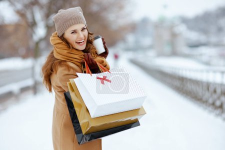 Téléchargez les photos : Femme élégante et souriante en chapeau marron et écharpe en manteau de chameau avec gants, sacs à provisions et tasse de boisson chaude à l'extérieur de la ville en hiver. - en image libre de droit
