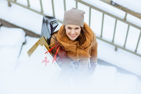 Téléchargez les photos : Vue supérieure d'une élégante femme souriante en chapeau marron et écharpe en manteau de chameau avec des sacs à provisions à l'extérieur de la ville en hiver. - en image libre de droit