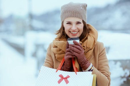Téléchargez les photos : Heureuse femme élégante en chapeau marron et écharpe en manteau de chameau avec gants, sacs à provisions et tasse de chocolat chaud à l'extérieur en ville en hiver. - en image libre de droit