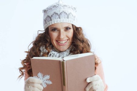 Photo for Hello winter. smiling trendy 40 years old woman in sweater, mittens, hat and scarf against white background with snowflake and book. - Royalty Free Image