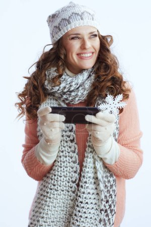 Photo for Hello winter. smiling stylish female in sweater, mittens, hat and scarf with snowflake sending text message using smartphone isolated on white. - Royalty Free Image