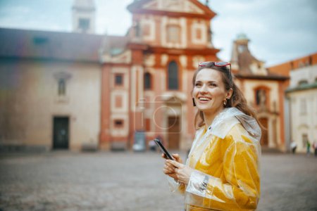 Téléchargez les photos : Heureuse femme touristique solo élégant en chemisier jaune et imperméable à Prague République tchèque en utilisant smartphone et la marche. - en image libre de droit