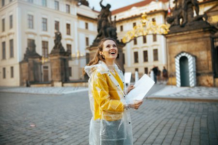 Photo for Smiling trendy middle aged traveller woman in yellow blouse and raincoat in Prague Czech Republic with map near Prague Castle. - Royalty Free Image