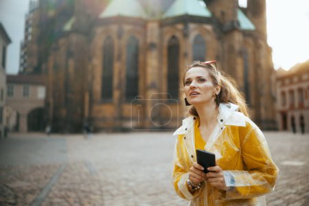 Foto de Sorprendió a la mujer viajera moderna en blusa amarilla y impermeable en Praga República Checa teniendo recorrido a pie y usando teléfono inteligente. - Imagen libre de derechos