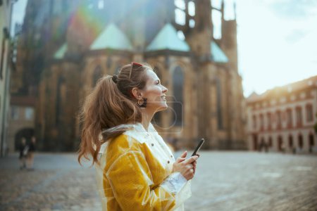 Photo for Surprised stylish woman in yellow blouse and raincoat in Prague Czech Republic enjoying promenade and using smartphone. - Royalty Free Image