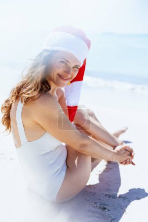Photo for Happy modern female in white swimwear with striped christmas hat sitting at the beach. - Royalty Free Image