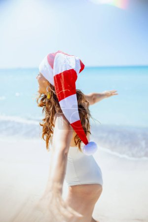 Foto de Mujer elegante feliz en traje de baño blanco con sombrero de Navidad rayado que se divierte en la playa. - Imagen libre de derechos