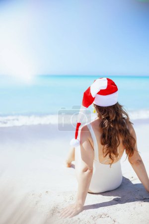 Foto de Visto desde atrás mujer en traje de baño blanco con el pelo ondulado largo y sombrero de Navidad rayado sentado en la playa. - Imagen libre de derechos