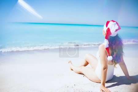Téléchargez les photos : Vu de derrière femelle moderne en vêtements de plage blancs avec de longs cheveux ondulés et chapeau de Noël rayé assis à la plage. - en image libre de droit