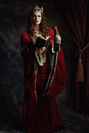 Téléchargez les photos : Portrait complet de la reine médiévale en robe rouge avec livre, bougie et couronne sur fond gris foncé. - en image libre de droit
