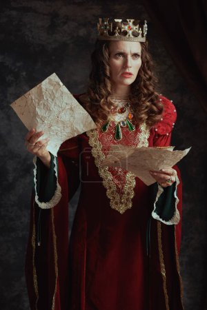 Photo for Unhappy medieval queen in red dress with parchment and crown on dark gray background. - Royalty Free Image