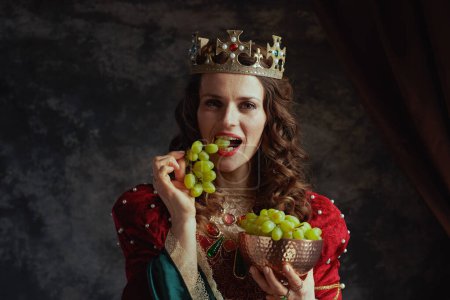 Photo for Medieval queen in red dress with plate of grapes and crown on dark gray background. - Royalty Free Image