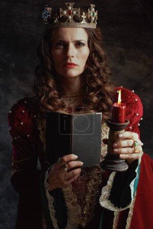 Photo for Medieval queen in red dress with book, candle and crown on dark gray background. - Royalty Free Image