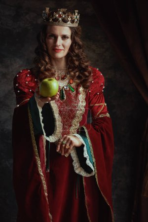 Téléchargez les photos : Reine médiévale en robe rouge avec pomme verte et couronne sur fond gris foncé. - en image libre de droit