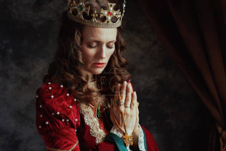 Foto de Reina medieval en vestido rojo con rosario y corona rezando sobre fondo gris oscuro. - Imagen libre de derechos