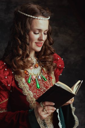 Foto de Sonriente reina medieval en vestido rojo con libro sobre fondo gris oscuro. - Imagen libre de derechos