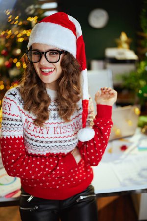 Photo for Christmas time. Portrait of happy elegant 40 years old business woman in santa hat in modern green office with Christmas tree in eyeglasses in red Christmas sweater. - Royalty Free Image