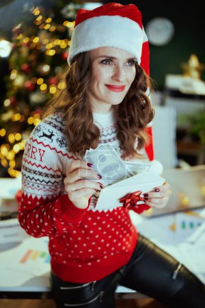 Photo for Christmas time. smiling modern middle aged small business owner woman in santa hat and red Christmas sweater with envelope and money in modern green office with Christmas tree. - Royalty Free Image