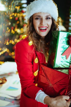 Téléchargez les photos : Noël. Portrait de heureuse élégante propriétaire de petite entreprise femme en santa chapeau et veste rouge avec des boîtes cadeaux dans le bureau vert moderne avec arbre de Noël. - en image libre de droit