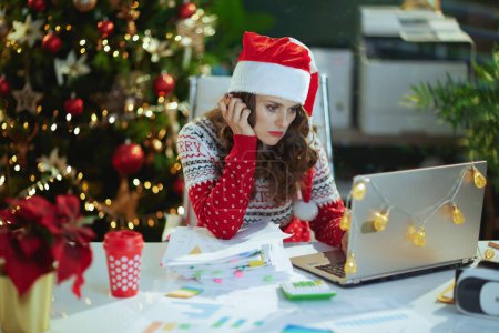 Photo for Christmas time. tired modern 40 years old business woman in santa hat and red Christmas sweater with documents and laptop working in modern green office with Christmas tree. - Royalty Free Image