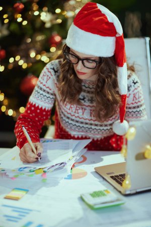 Photo for Christmas time. elegant small business owner woman in santa hat in eyeglasses in red Christmas sweater with documents and laptop working in modern green office with Christmas tree. - Royalty Free Image