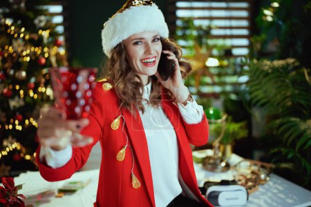 Téléchargez les photos : Noël. femme d'affaires élégante et souriante en santa chapeau et veste rouge avec verre à vin parlant sur un smartphone dans un bureau vert moderne avec arbre de Noël. - en image libre de droit