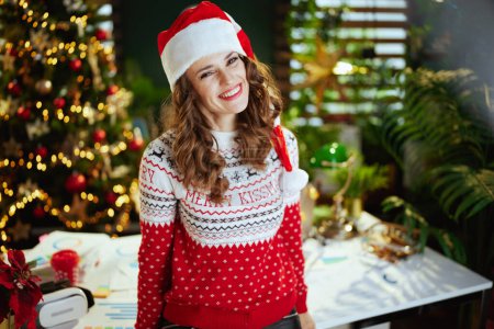 Téléchargez les photos : Noël. Portrait de femme d'affaires souriante moderne de 40 ans en chapeau de Père Noël dans un bureau vert moderne avec arbre de Noël en pull de Noël rouge. - en image libre de droit