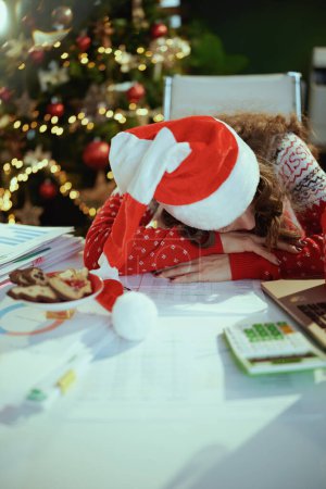 Foto de En Navidad. mujer de negocios elegante cansado en sombrero de santa y suéter rojo de Navidad con documentos que duermen en la oficina verde moderna con árbol de Navidad. - Imagen libre de derechos