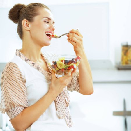 Photo for Happy young housewife eating fresh fruit salad in kitchen - Royalty Free Image