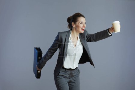 Photo for Smiling modern female employee in gray suit with coffee cup and briefcase running isolated on gray background. - Royalty Free Image