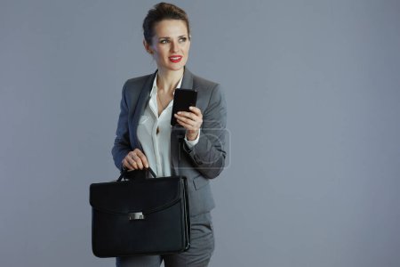 Téléchargez les photos : Chère femme d'affaires moderne en costume gris avec smartphone et mallette sur fond gris. - en image libre de droit