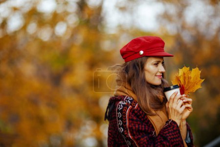 Foto de Hola otoño. mujer de 40 años pensativa en sombrero rojo con hojas de otoño y bufanda en el parque de la ciudad. - Imagen libre de derechos