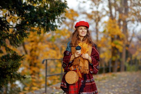 Photo for Hello autumn. smiling stylish 40 years old woman in red hat with autumn leafs, scarf, bag and coffee walking in the city park. - Royalty Free Image