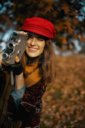 Téléchargez les photos : Bonjour l'automne. heureuse femme élégante en chapeau rouge avec caméra vidéo rétro, écharpe et gants dans le parc de la ville. - en image libre de droit