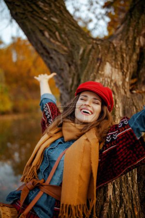 Photo for Hello autumn. happy young female in jeans shirt and red hat with scarf near tree in the city park. - Royalty Free Image