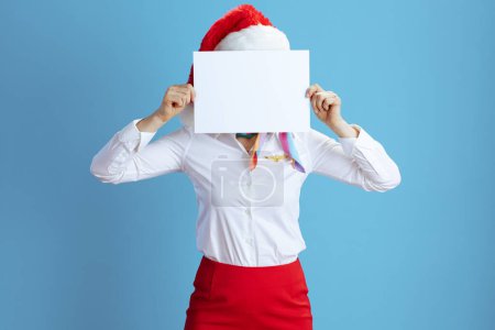 Photo for Modern flight attendant woman isolated on blue background in uniform with Santa hat showing blank a4 paper sheet. - Royalty Free Image