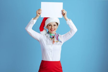Téléchargez les photos : Hôtesse de l'air féminine moderne souriante isolée sur fond bleu en uniforme avec chapeau de Père Noël montrant feuille de papier blanc a4. - en image libre de droit