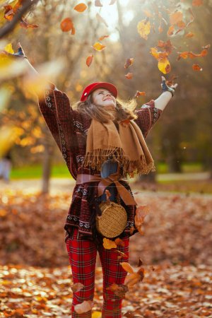 Foto de Hola otoño. sonriente elegante hembra en sombrero rojo con bufanda, guantes y bolsa lanzando hojas de otoño en el parque de la ciudad. - Imagen libre de derechos