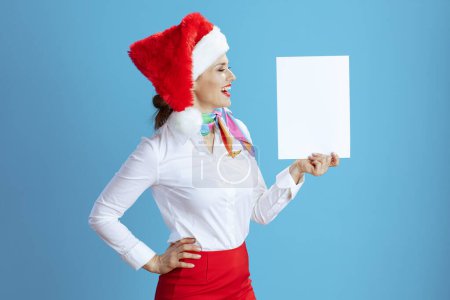 Téléchargez les photos : Souriant hôtesse de l'air féminine élégante sur fond bleu en uniforme avec chapeau Santa montrant feuille de papier blanc a4. - en image libre de droit