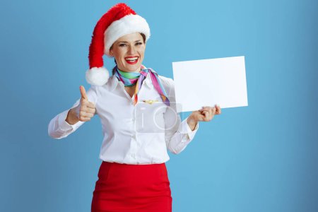 Téléchargez les photos : Heureuse hôtesse de l'air féminine moderne sur fond bleu en uniforme avec chapeau Santa montrant feuille de papier blanc a4. - en image libre de droit