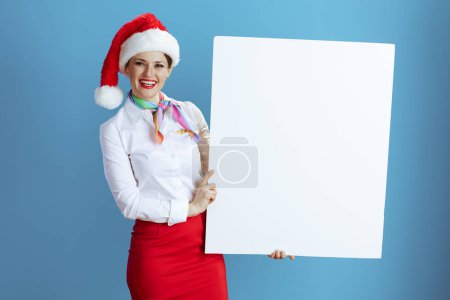 Foto de Sonriente elegante azafata de vuelo femenino sobre fondo azul en uniforme con sombrero de Santa mostrando tablero en blanco. - Imagen libre de derechos