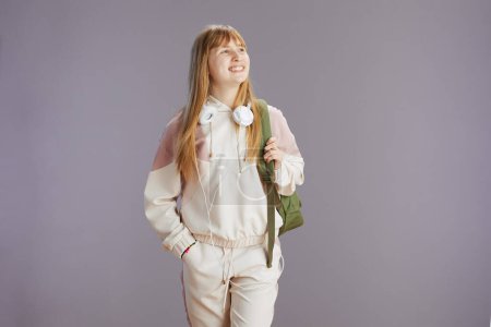 Photo for Portrait of smiling modern teenager girl in beige tracksuit with backpack and headphones isolated on grey background. - Royalty Free Image