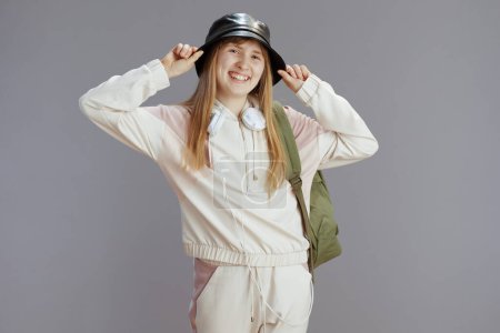 Foto de Retrato de una adolescente moderna sonriente en chándal beige con mochila, auriculares y sombrero aislados sobre fondo gris. - Imagen libre de derechos