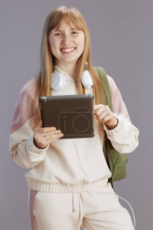 Foto de Chica de moda feliz en chándal beige con mochila, auriculares y tableta digital aislada en gris. - Imagen libre de derechos